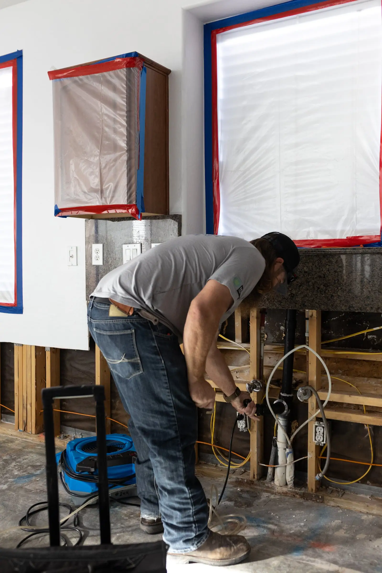 Fast Track technician working on exposed wall framing and plumbing after wet drywall was removed, with the cabinets sealed in plastic